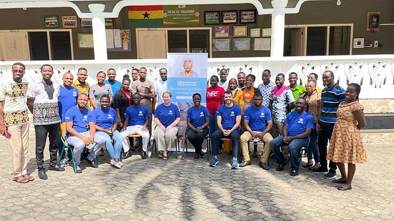 Participants, mentors and WHO staff members are in 2 rows, 1 sitting and 1 standing, around a WHO banner advertising the training. They are looking at the camera and smiling